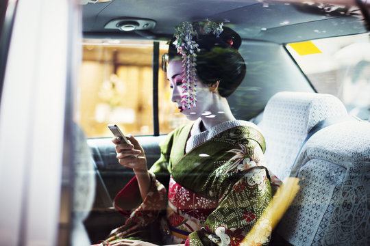 A woman dressed in the traditional geisha style, wearing a kimono and obi, with an elaborate hairstyle and floral hair clips, with white face makeup with bright red lips and dark eyes in a car using a smart phone. 