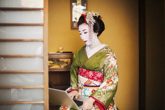 A woman dressed in the traditional geisha style, wearing a kimono and obi, with an elaborate hairstyle and floral hair clips, with white face makeup with bright red lips and dark eyes using a laptop computer. 