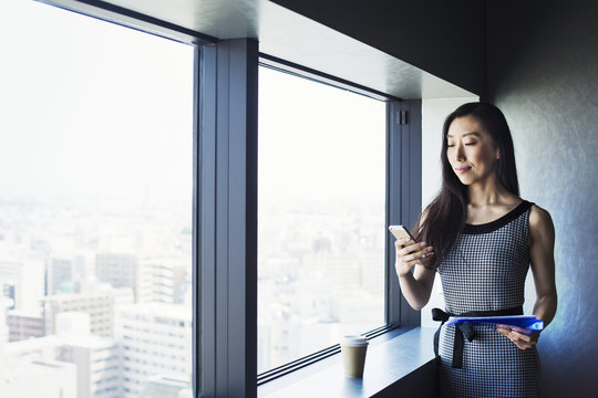 A Business Woman By A Window With A View Over The City, Using Her Smart Phone. 