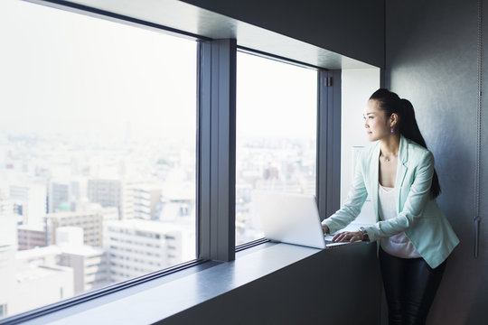 A business woman by a window with a view over the city, looking out. Laptop. 