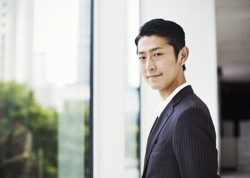 A businessman in the office, by a large window, looking at the camera. 