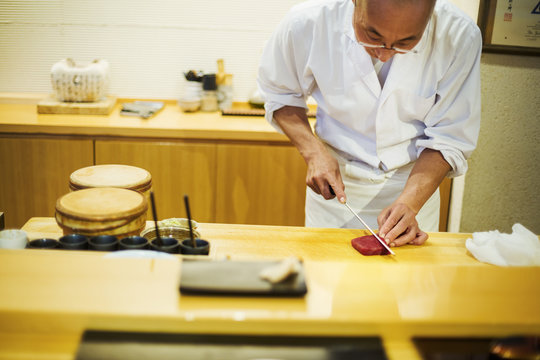 A chef working in a small commercial kitchen, an itamae or master chef slicing fish with a large knife for making sushi