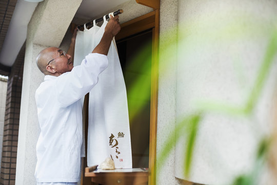 A chef in a small commercial kitchen, an itamae or master chef drawing a curtain across a door. 
