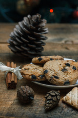 Chocolate chip cookies on rustic background. Nearby lie cinnamon sticks and cones.Behind is tree with toys and garlands. New Year holiday.
