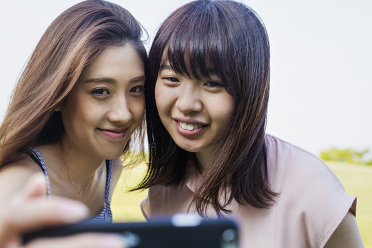 Two smiling young women with long brown hair, holding a mobile phone, taking selfie.
