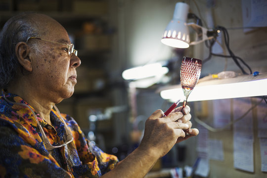 A senior craftsman at work in a glass maker's studio workshop, in inspecting red wine glass with cut glass decoration against the light. 