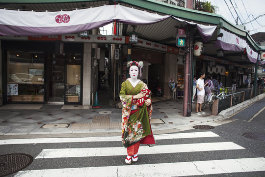 A woman dressed in the traditional geisha style, wearing a kimono and obi, with an elaborate hairstyle and floral hair clips, with white face makeup with bright red lips and dark eyes crossing a street. 
