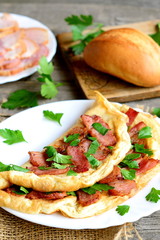 Homemade bacon omelet. Delicious omelet stuffed with bacon and parsley on a plate, bacon slices on a plate, bread roll, fork, knife, cutting board, fresh parsley sprigs on old wooden table. Closeup