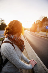 Thoughtful woman traveler with a backpack walking along the road. Red-haired woman in a sweater. Sunset. Highway.