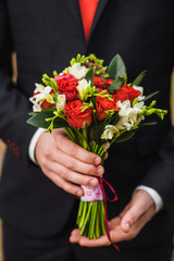 Beautiful bouquet of fresh flowers in hands of anonymous elegant man dressed in modern black suit, white shirt, red necktie. Close up color vertical image of male hands holding bunch of bright flowers