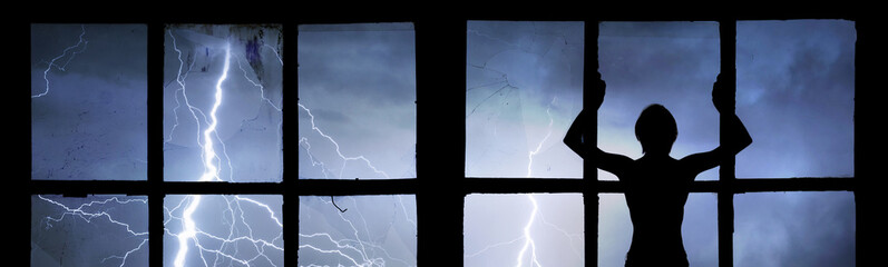 Silhouette of man watching lightning, thunder, rain and storm through broken window of old...