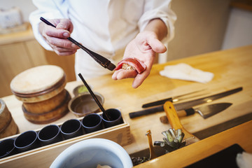 A chef working in a small commercial kitchen, an itamae or master chef making sushi, pasting sauce on fish. 