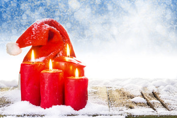 Weihnachtsmütze und 4 brennende Kerzen auf verschneitem Holzboden vor blauem Hintergrund mit Bokeh...