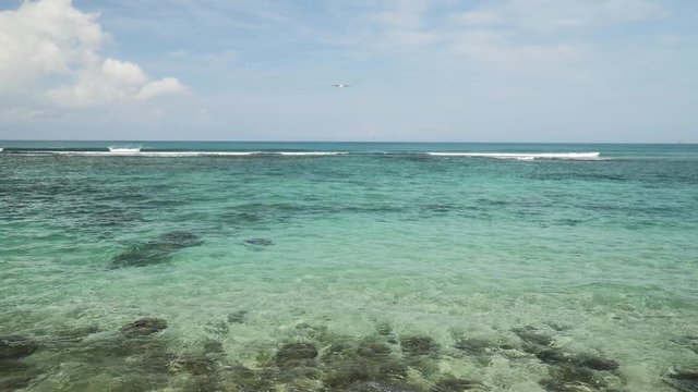 Airplane landing on island Bali airport under blue sea with waves. Cinematic shot of overhead flying aircraft with shadow on beautiful ocean on cloudy sky background. Focal length 50 mm. Slow motion