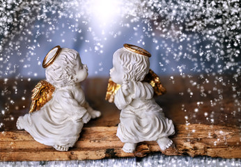 Zwei Engel sitzen einander zugewandt auf Holz, um sie herum fällt Schnee