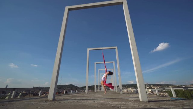 Beautiful dancer on aerial silk. Young sexy woman performs acrobatic stunts on the rooftop against sky with clouds