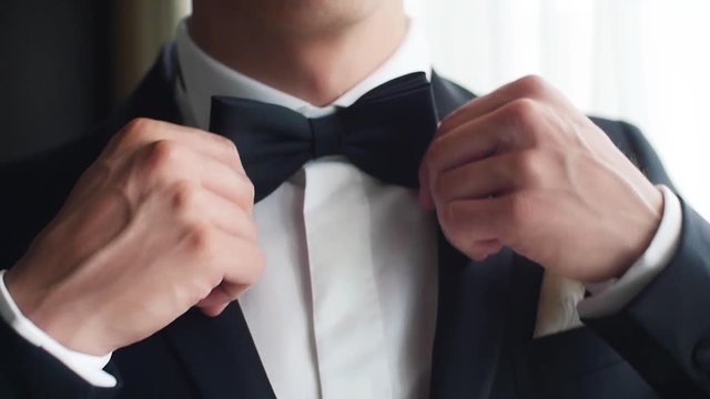 Man adjusts black bow tie closeup slow motion. Male hands correct bowtie on white shirt wear suit prepare for wedding. Gentleman fashion trends fashionable stylish businessman outfit party light image