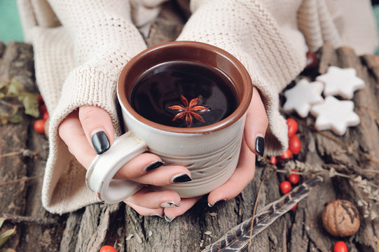 Female hands in warm clothes holding a ceramic cup of hot tea on a wood bark with some rose hips fruits