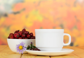 Bowl dog rose berries, cup tea on background autumn leaves.