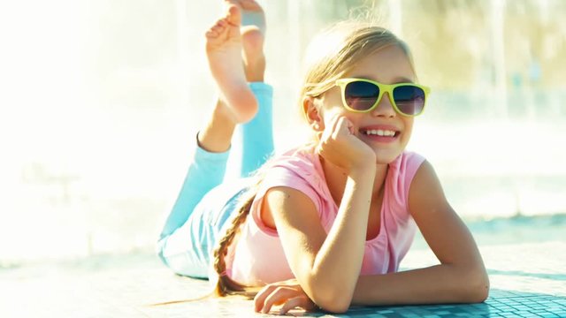 Portrait child lying on fountain background at sunny day and waving hand. Zooming