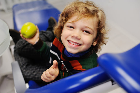 curly-haired child in the dental office smiling and holding a green apple