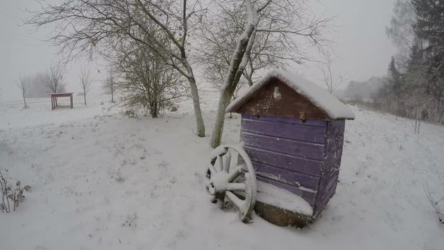 Snow falling on violet beehive with wooden wheel in countryside yard, time lapse 4K 