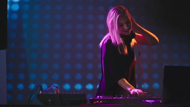 PAN with slow motion of beautiful blond DJ woman dancing behind mixer, LED video wall in background