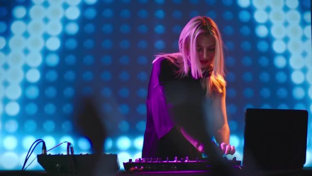 Slow motion of young blond DJ woman dancing behind mixing console at party in nightclub, LED video wall in background