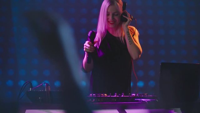 Tilt up of female blond DJ jumping and dancing behind decks in nightclub, LED video wall in background