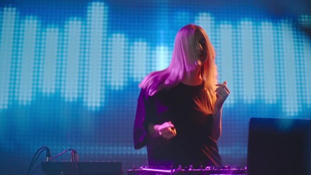 Young blond female DJ dancing excitedly behind decks in nightclub