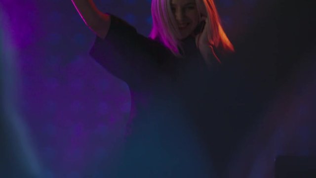 Tilt up of blond female DJ jumping excitedly and playing decks on party in nightclub
