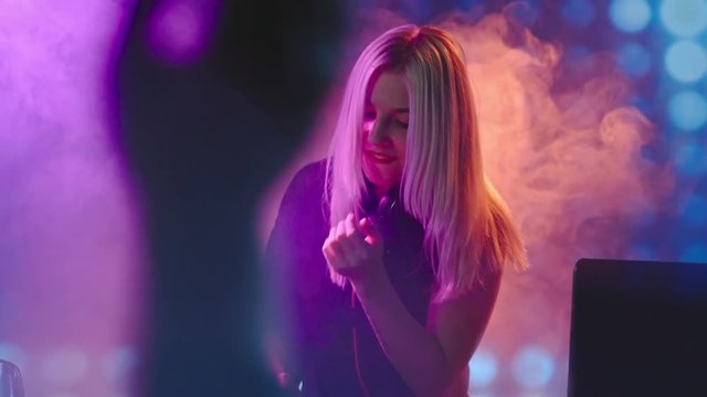 Tilt up of seductive female DJ with blond hair dancing behind decks and singing at party in nightclub 
