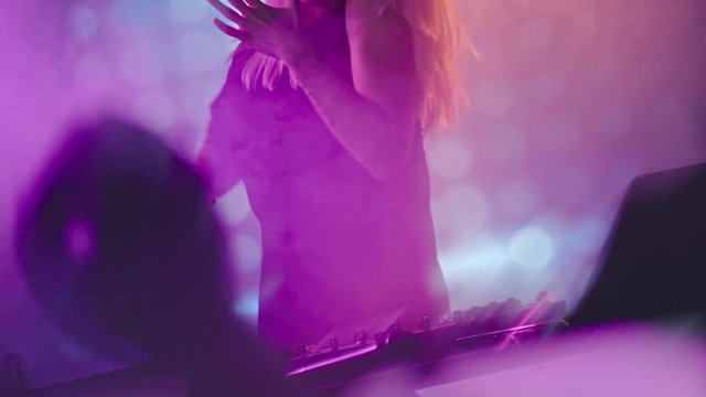 Dutch angle of blond female DJ dancing and playing music on decks at smoky nightclub 