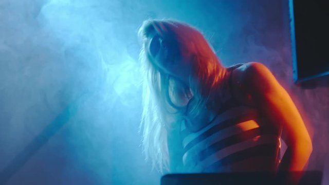 Young blond woman dancing behind DJ mixer console in dark smoky nightclub