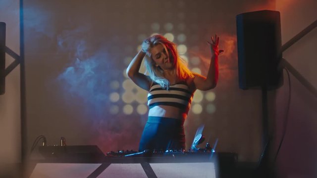 Young woman seductively dancing behind mixer console and DJing in nightclub