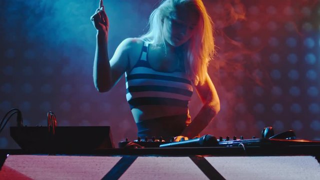 Slow motion of blond sexy woman DJ dancing and singing behind decks on party in dark smoky club