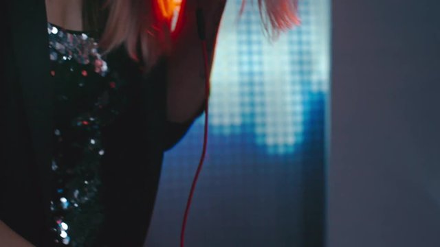 Tilt down of beautiful blond woman in sparkling dress DJing in dark nightclub 