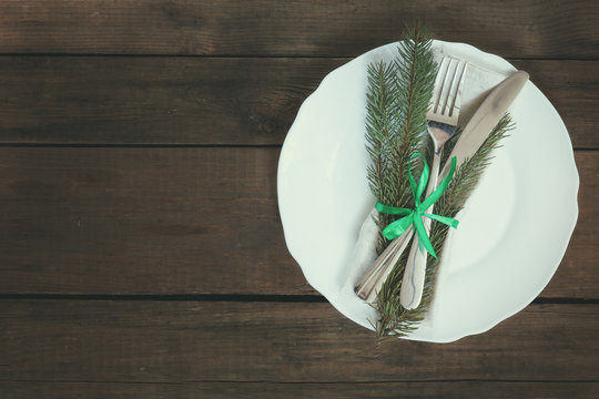 Country style christmas decoration. White empty plate with fir tree, fork and knife tied with a green ribbon on old wooden brown background.