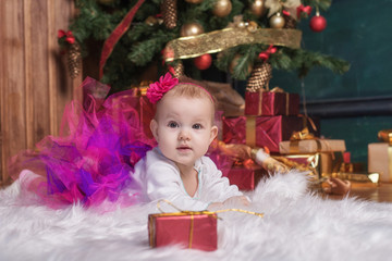 Cute baby girl wearing pink skirt and red headband, lying on white carpet near christmas trees. Christmas gifts.