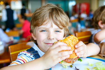 Cute healthy preschool boy eats hamburger sitting in cafe outdoors