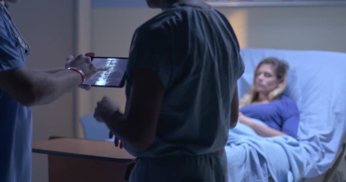 Young woman rests in hospital bed while medical staff look at X rays on tablet computer in foreground.  Focus on X rays, with patient to side of screen.  Evening lighting.