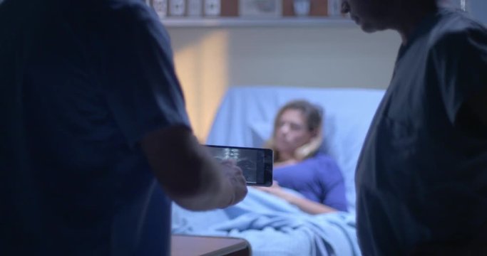 Young woman rests in hospital bed while medical staff look at X rays on tablet computer in foreground.  Focus on patient to start, with rack focus to X rays after.  Evening lighting.