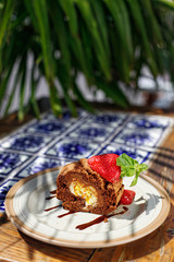 Brownie roll-Mexican Chocolate and Fruit cake, visible Strawberries And Mint Leaves, sunlit and vibrant With blurred Palm Leaves In The Background, vertical 