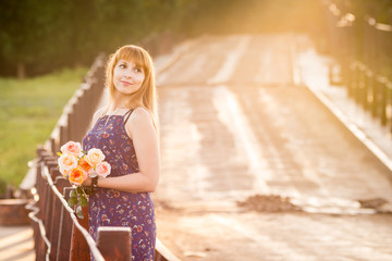 The charming young girl standing on a rustic bridge at dawn sun with a bouquet of roses