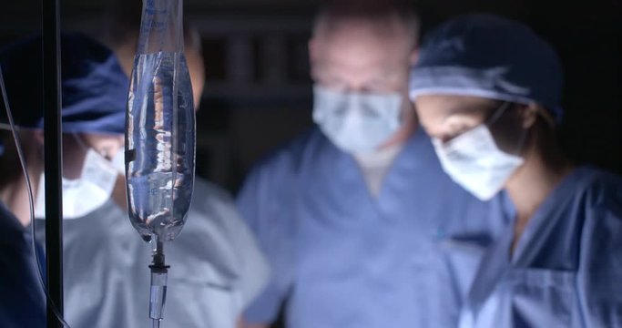 Surgical team of doctors and nurses in scrubs and masks, working in background with saline drip bag in focus in foreground.