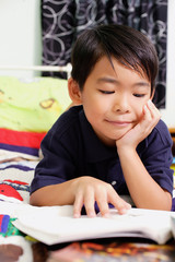 Boy lying on bed, hand on chin, reading a book