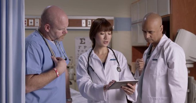 Male nurse and two doctors look at information on tablet computer and check an X-ray photo in hospital exam room.  Medium shot.