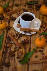 Obraz na płótnie Canvas Small white cup of coffee, cinnamon sticks, cocoa beans, star anise, hazelnuts and mandarins on wooden background