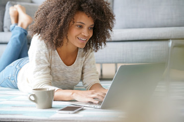Cheerful mixed-race woman websurfing with laptop, laid on carpet