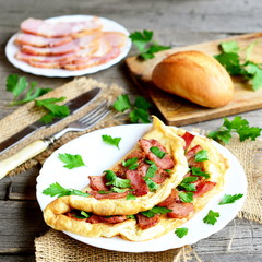 Delicious bacon omelette. Omelette stuffed with bacon and parsley on a plate, bacon slices on a plate, bread roll, fork, knife, chopping board, fresh parsley on old wooden background. Homemade cuisine